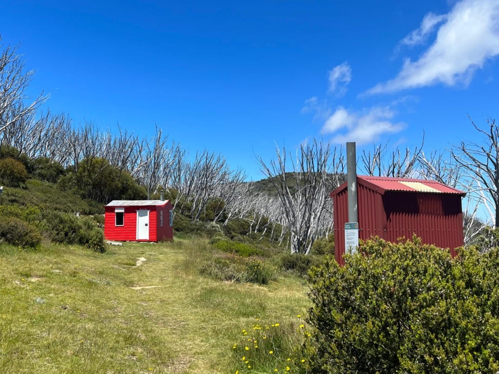 A red hut with a matching red drop toilet on one side, set against a clear blue sky